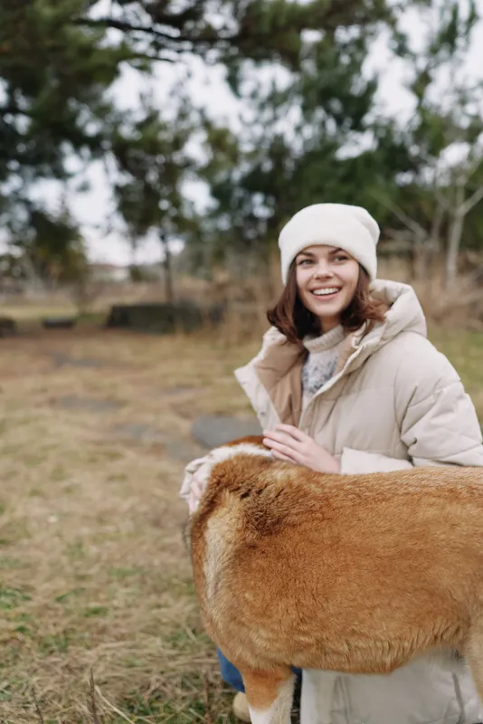 Woman in a warm beige coat and knit hat smiles while hugging a large fluffy animal in an open field during chilly weather, creating a cozy rural moment for a rustic lifestyle shot.