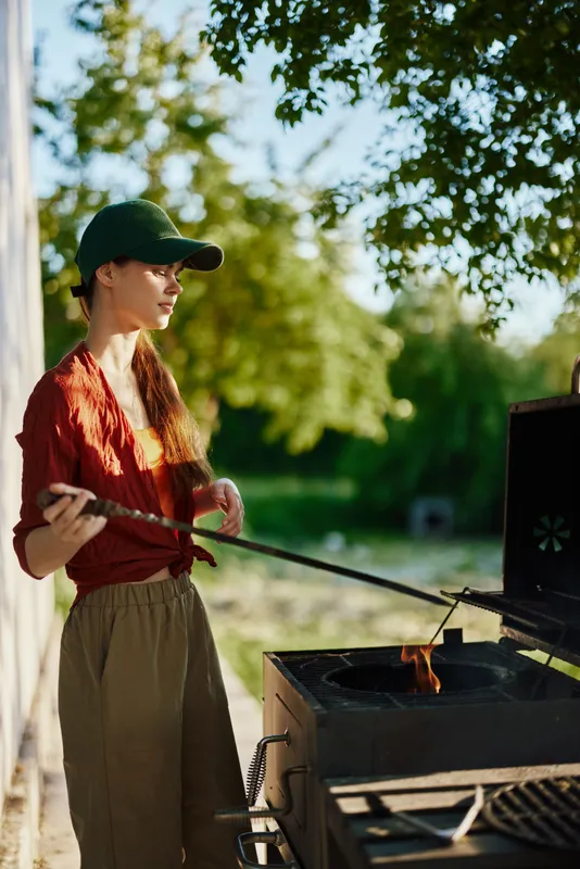 Young woman grilling outdoors in a sunny garden, wearing a casual outfit and a cap, showcasing joy and relaxation during summer cooking moments