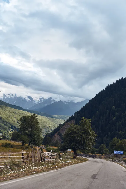 Mountain road winds through a valley with trees and rugged hills beneath cloudy skies, capturing a quiet rural landscape ideal for travel, nature, and outdoor photography.