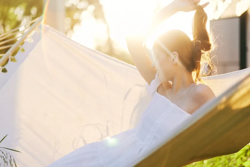 Young woman with light brown hair enjoying a sunny day in a white dress on a hammock, radiating relaxation and happiness amidst a lush green background