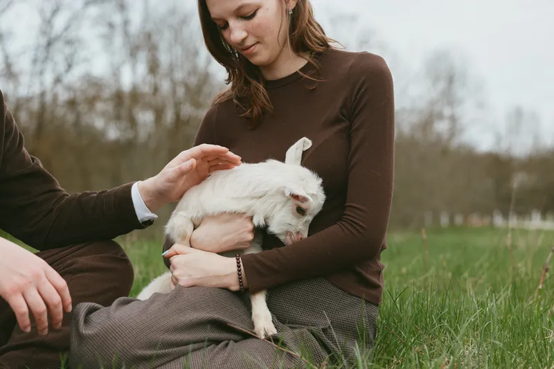 Outdoor lifestyle moment with a woman gently cradling a goat kid in a grassy field, warm natural light, calm interaction, pastoral mood, soft colors, serene countryside atmosphere