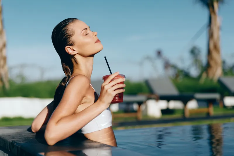 Cheerful outdoor moment with a woman by a pool, sunlight on her skin, relaxation in a sunlit setting, savoring a refreshing beverage with a gentle breeze and blue water in the background