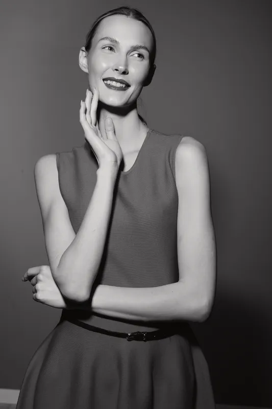 Tall slender white woman with pale skin smiles thoughtfully, wearing sleeveless dress against dark background in black and white portrait photography.