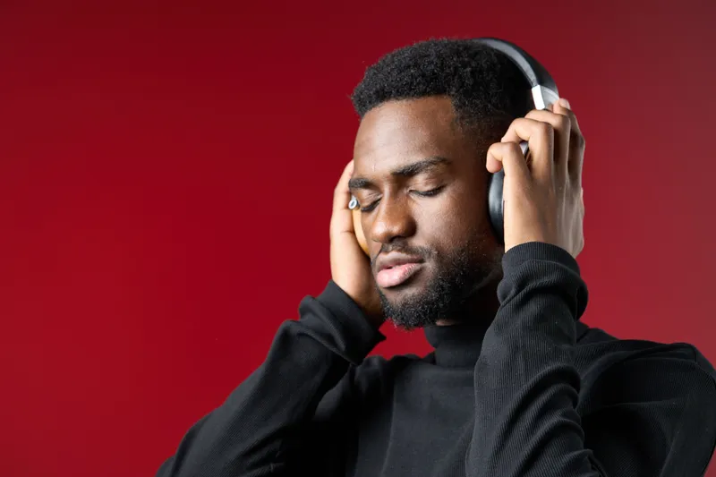 A young Black man in a black turtleneck listens to music with closed eyes, enjoying the rhythm against a vibrant red background, reflecting emotions of relaxation and focus