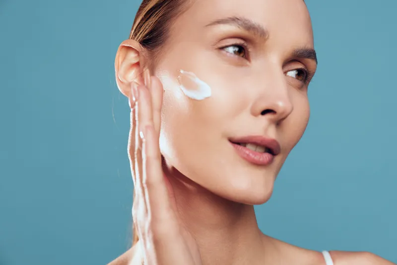 Young woman applies white cream to her cheek in a close portrait against a blue background, conveying skincare, wellness, and self care with soft studio lighting.