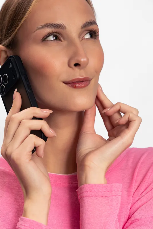 Young woman in pink sweater holding smartphone to ear, looking away thoughtfully. Studio portrait on white background. Concept for communication, technology, and lifestyle.