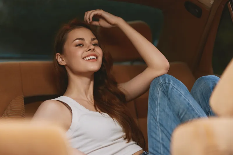 Smiling young woman in casual wear sitting in car, expressing joy and relaxation, with denim jeans and a simple white tank top, creating a warm and inviting atmosphere.