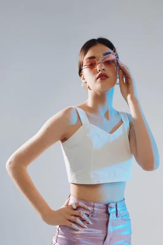 Young woman beauty fashion model wearing pink sunglasses and white crop top poses confidently with hand on hip. Studio shot with soft lighting on gray background.
