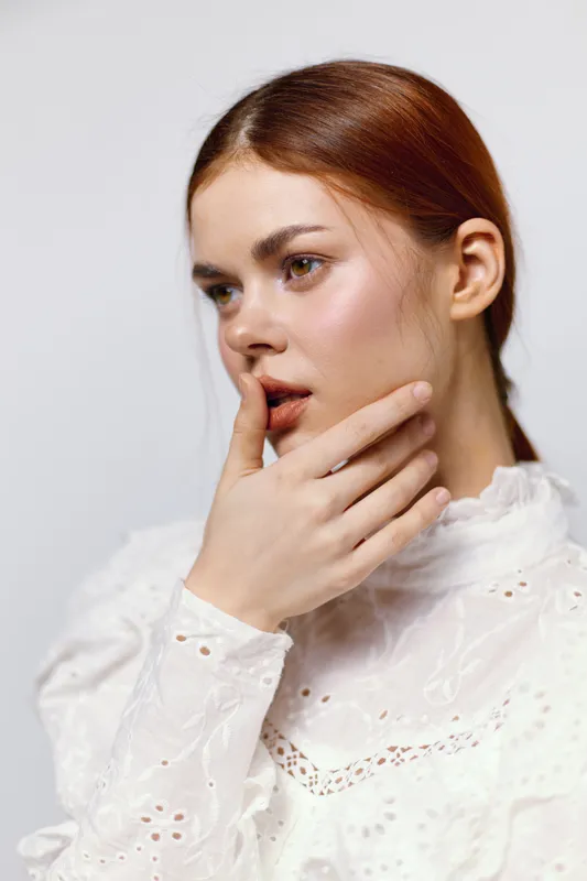 Young woman in a white lace blouse contemplating emotions with a soft expression on a neutral background, ideal for fashion or beauty concepts