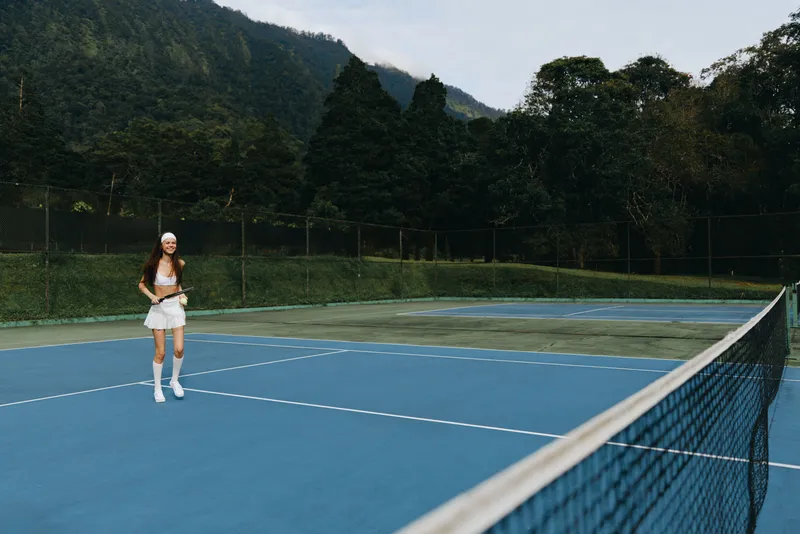 Young female tennis player smiling on a blue court surrounded by lush greenery, showcasing an active lifestyle and passion for sports