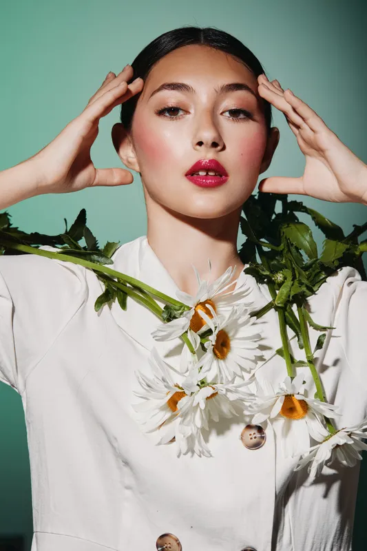 Young woman in white fashion outfit with vibrant daisy flowers around her neck poses confidently. Glamorous makeup and clean background highlight the beauty concept.
