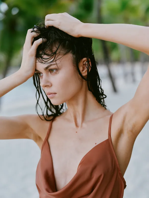 Portrait of a young woman on a sunlit beach, wet hair swept back, wearing a rust swimsuit, conveying vulnerability and quiet strength in a candid, intimate frame.