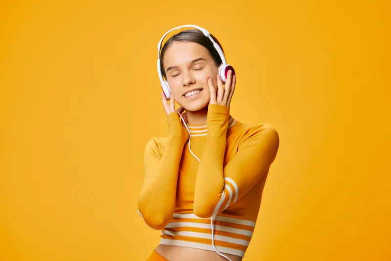 Female fitness enthusiast wearing orange activewear, listening to music with headphones, expressing energy and focus in a bright studio backdrop.