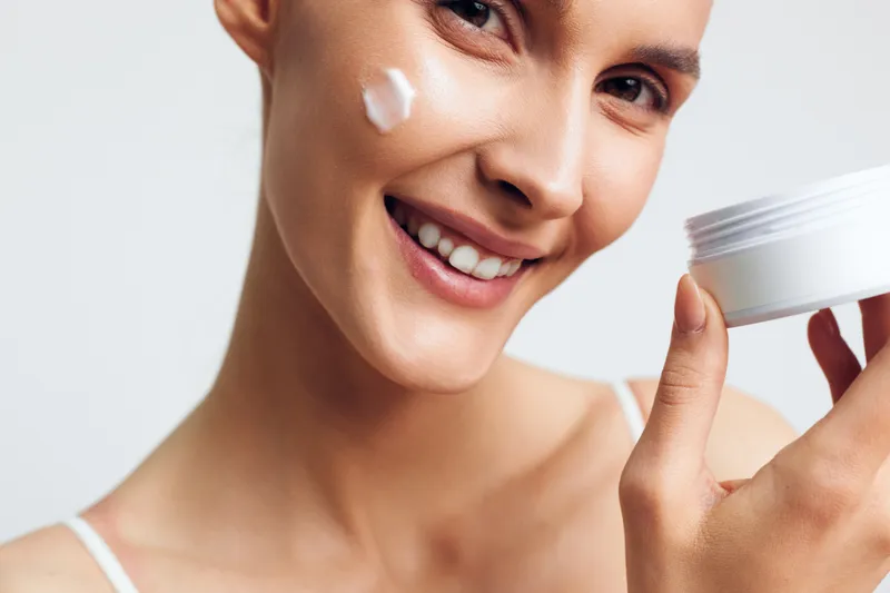 A smiling young woman applies white cream on her cheek while holding an open jar of skincare product in a bright studio. Concept for beauty, skin care, and hydration.