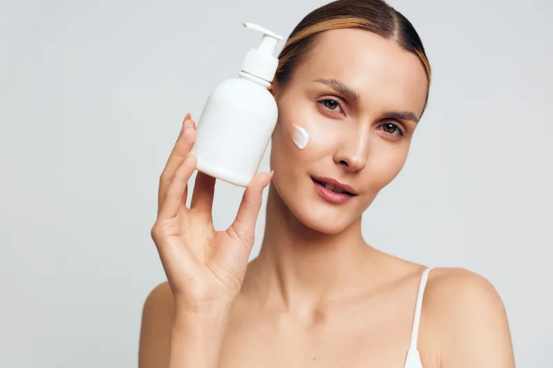 Young woman with clear skin holds white lotion bottle near face with cream dot on cheek shows skincare product for beauty routine isolated on light background.