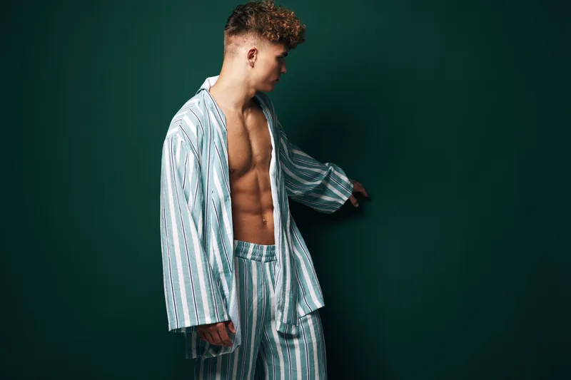 Young man in striped pajamas standing against dark background looking to side with bare chest and relaxed posture in casual homewear for comfort and style.
