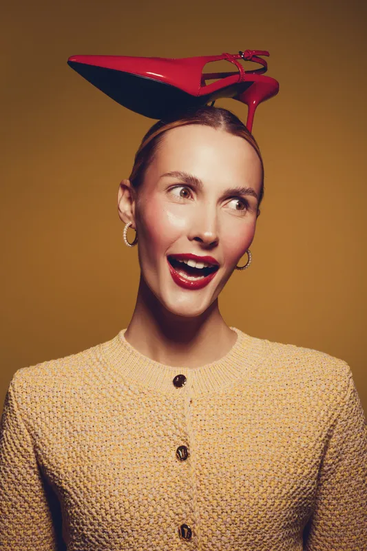 Young woman with red high heel shoe on head smiles and looks aside. Fashion concept with playful mood and creative styling on yellow background in studio lighting.