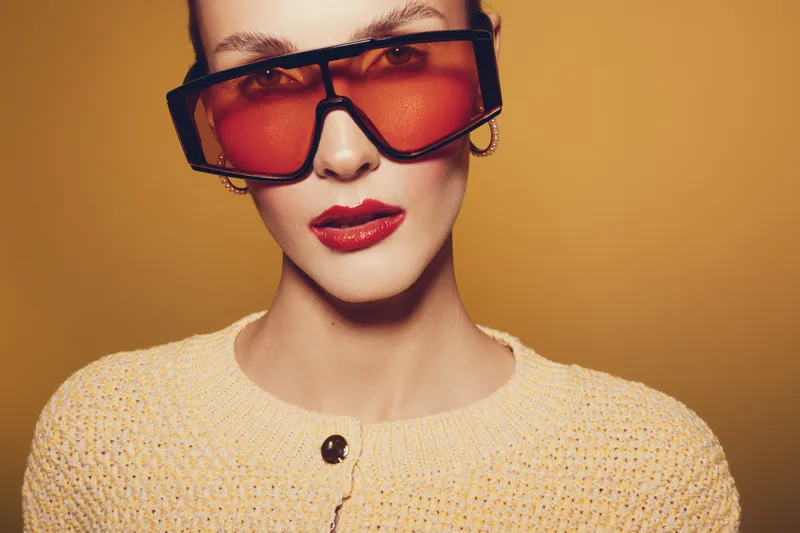 Stylish woman with red lipstick and oversized orange sunglasses poses against yellow background. She wears a knitted beige sweater and gold hoop earrings. Concept of fashion and modern style.