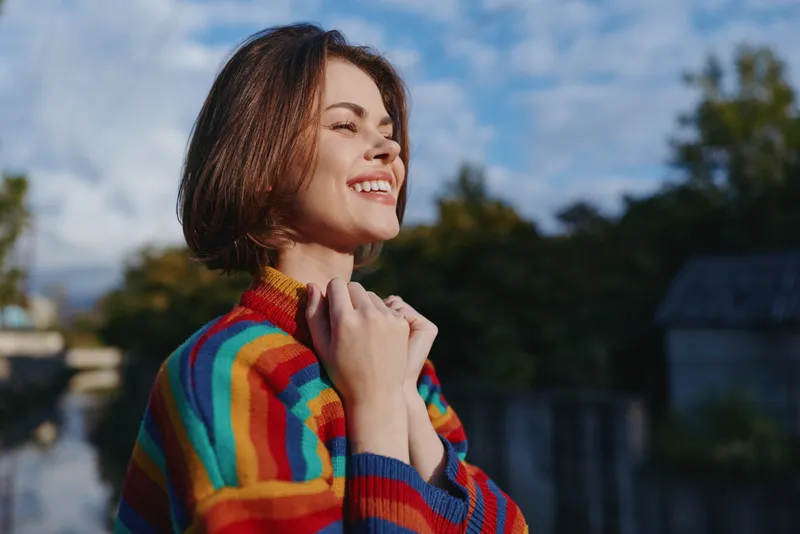 woman smiling striped sweater outdoor short hair looking up casual outfit autumn light hands near chest; mid-30s model with short bob haircut, joyful expression, colorful knit by riverside in morning