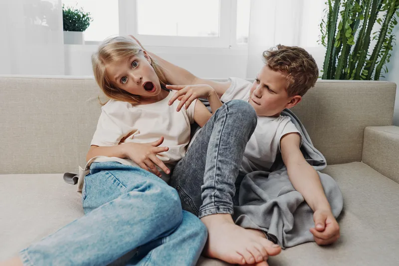 Two children sitting on sofa, boy playfully pushing girl with foot, girl showing surprised face, casual home interior with bright natural light.