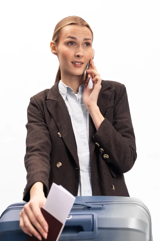 Young woman in brown blazer talking on phone while holding passport and boarding pass near suitcase. Concept of travel, business trip, communication, and preparation for flight.