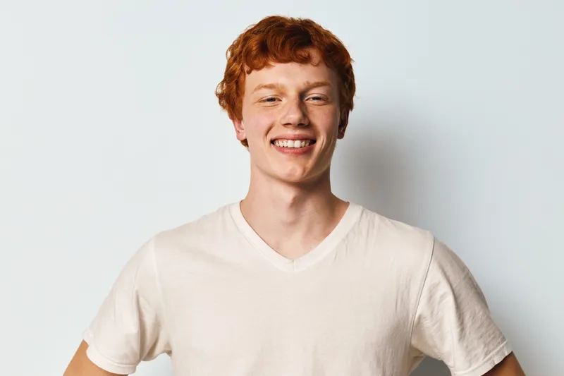 Young man with red hair smiling and posing with hands on hips in casual white t shirt on plain background. Portrait, happy and confident male adult indoors.