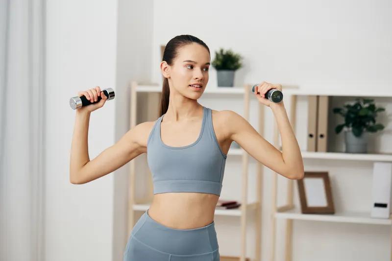 Fitness oriented woman in a light blue athletic set raises dumbbells for a shoulder press in a clean home gym corner, shelves and plants creating a calm studio vibe.