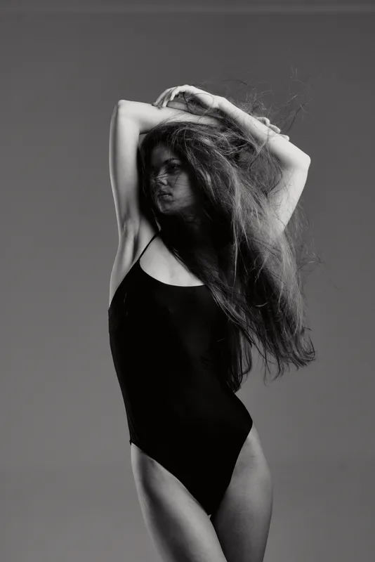 Black and white portrait of a woman in a black bodysuit posing with arms raised and long hair flowing, artistic and stylish.