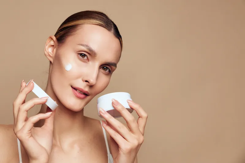 Young woman with clear skin applying white face cream on cheek. Close-up portrait on beige background. Concept of skincare, beauty, wellness, and self-care with soft lighting.