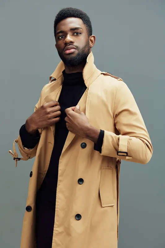 Stylish young African man in a beige trench coat, exuding confidence with a slight smile against a muted gray background, perfect for fashion and lifestyle themes