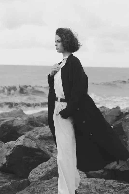 Woman standing on rocky coast in flowing black coat and white trousers, sea breeze enhances fashion and elegance in a monochrome outdoor scene with bold horizon and dramatic mood.