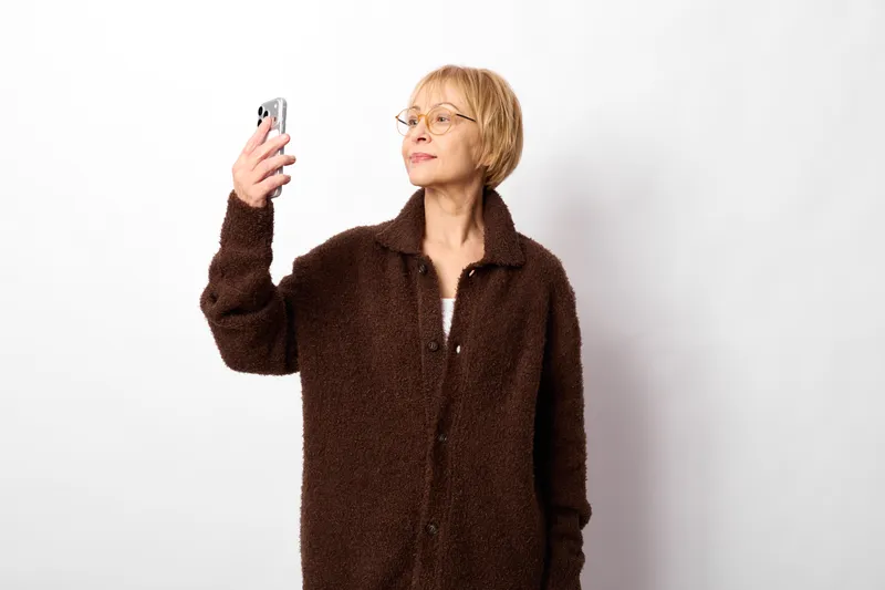 Woman holding smartphone and wearing glasses, portrait of mature adult with neutral smile, communicating with technology, standing against white background, studio lighting.