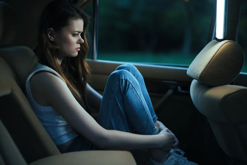 A contemplative young woman in casual attire sits in the back of a car, expressing emotions of solitude and introspection in a dimly lit environment.