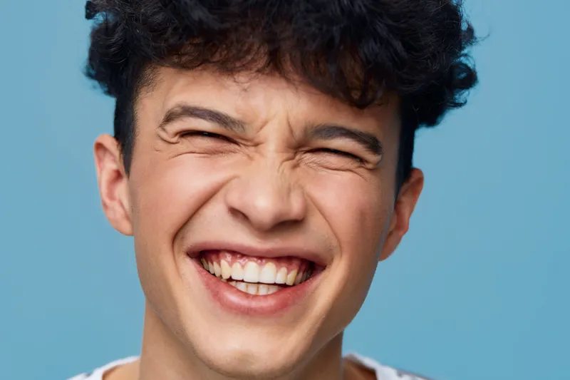 smiling man with curly hair showing joyful expression, close-up portrait with blue background, natural light capturing happiness and youthfulness.