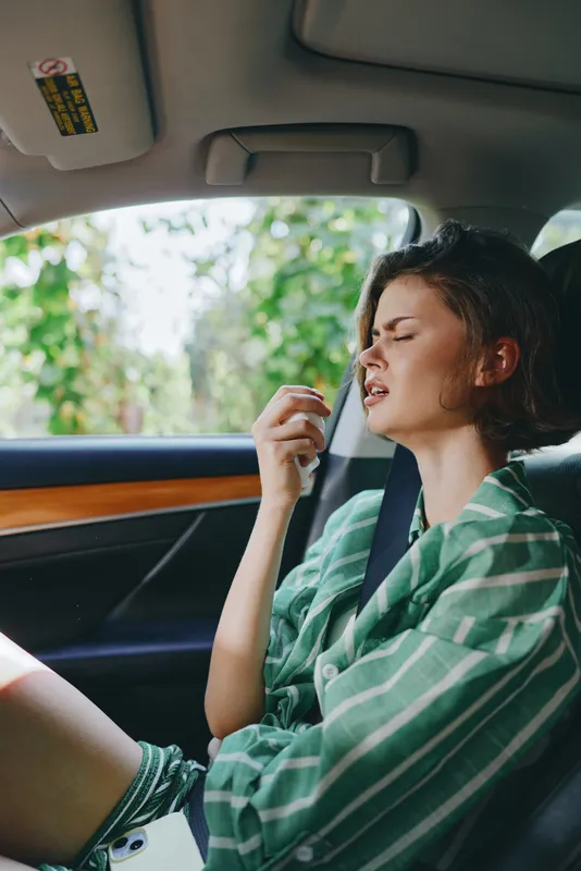Young woman sits in a car wearing a green striped shirt, eyes closed, and a relaxed pose as she rests a hand near her mouth.