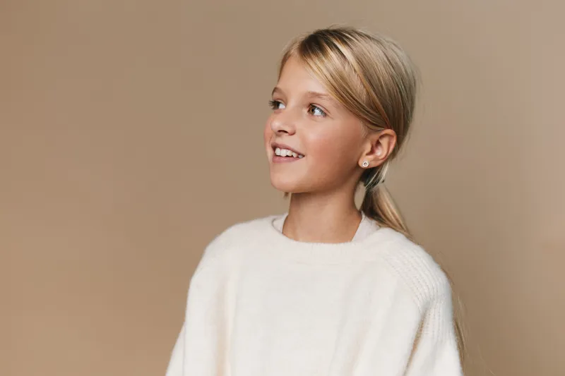 A young girl smiles softly while looking to the side, wearing a cozy white sweater with a neat ponytail, set against a warm beige background and soft lighting.