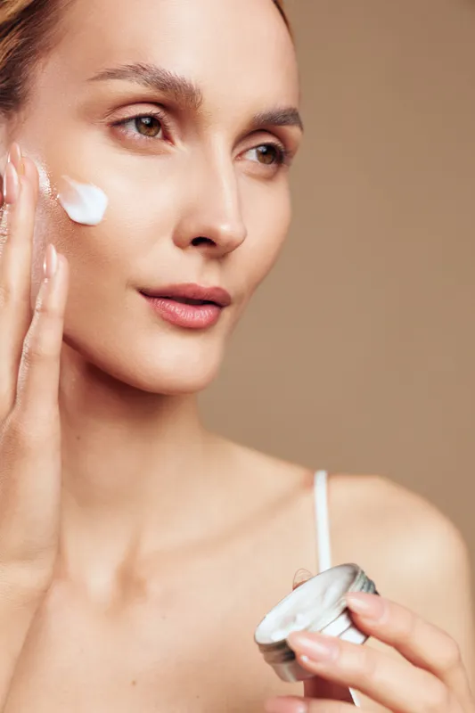Woman skincare beauty, applying cream to face with gentle touch, moisturizing in studio light, holding cream product, calm expression, neutral background, care routine