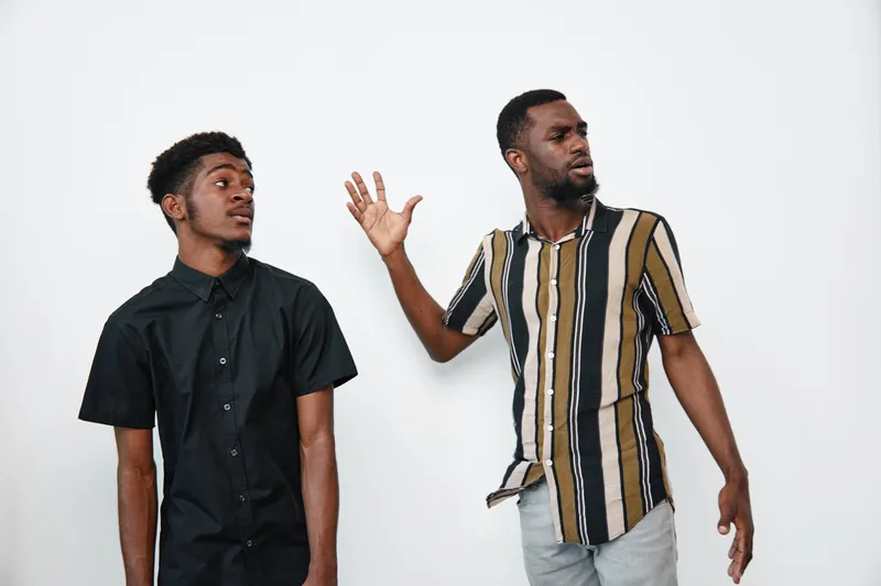 Two African American men with dark skin standing against white background, one wearing black shirt and the other striped shirt, engaged in conversation with expressive gestures.