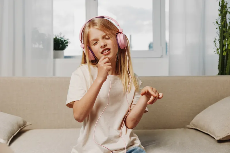 Young girl with long blonde hair wearing pink headphones enjoys music passionately while sitting on beige sofa in bright living room with natural light and casual outfit.