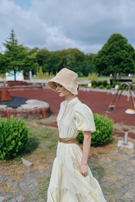 Woman in a light cream dress and straw hat walks through a park setting, calm and graceful, enjoying a sunny outdoor moment under blue skies.