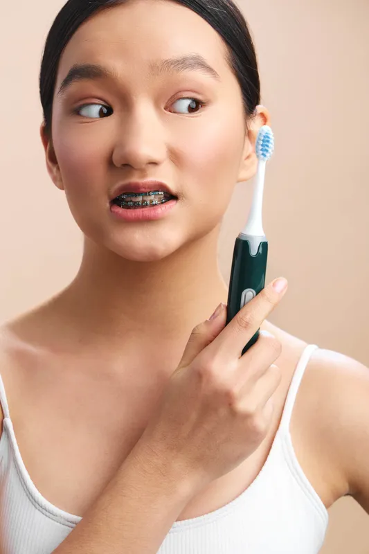 asian woman with braces is brushing her teeth with an electric toothbrush in a bright, minimal setting, presenting a fresh, healthy dental care moment for daily routine.