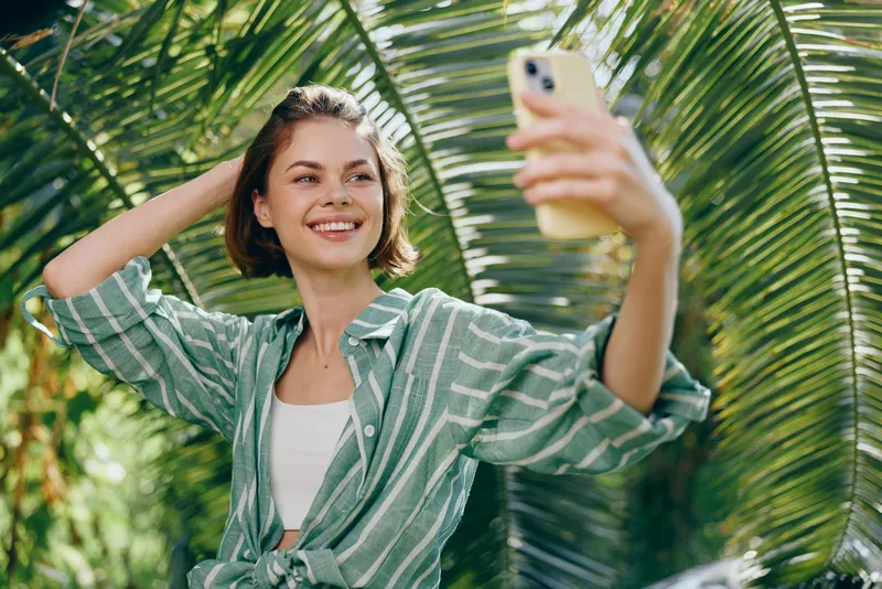 Smiling young woman takes a sunny selfie among lush palm fronds, wearing a striped green shirt.