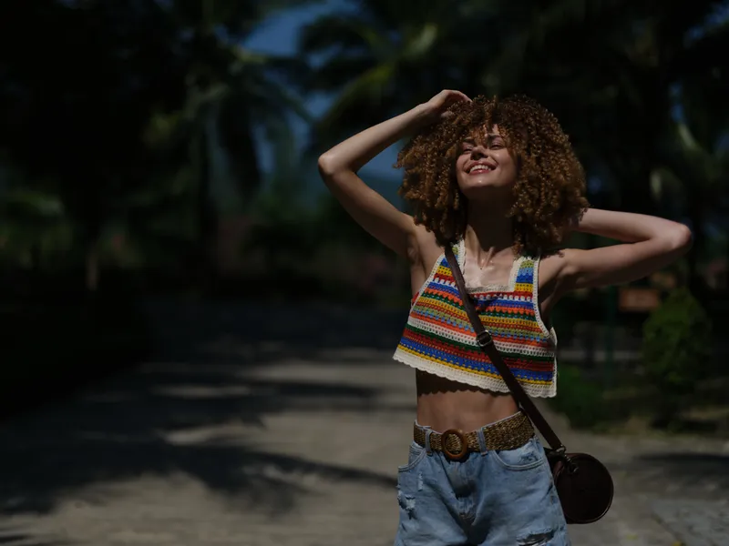 Smiling woman enjoying summer outdoors in colorful crochet top and denim shorts with curly hair and relaxed pose under natural sunlight.