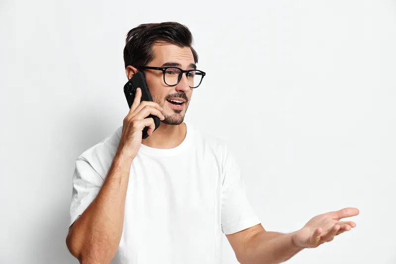 Casual young man talking on mobile phone while gesturing with hand, wearing white t-shirt and glasses, standing against solid white background. Communication concept.
