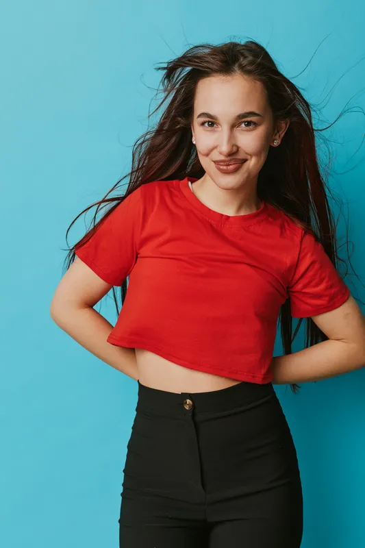 Young woman with long hair wearing red crop top and black pants posing with hands on hips against blue background smiling confidently at camera in studio portrait.