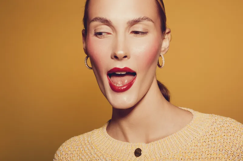 Young woman with red lipstick and hoop earrings looks sideways with open mouth on orange background. Concept of beauty, fashion, makeup, and female portrait in studio lighting.