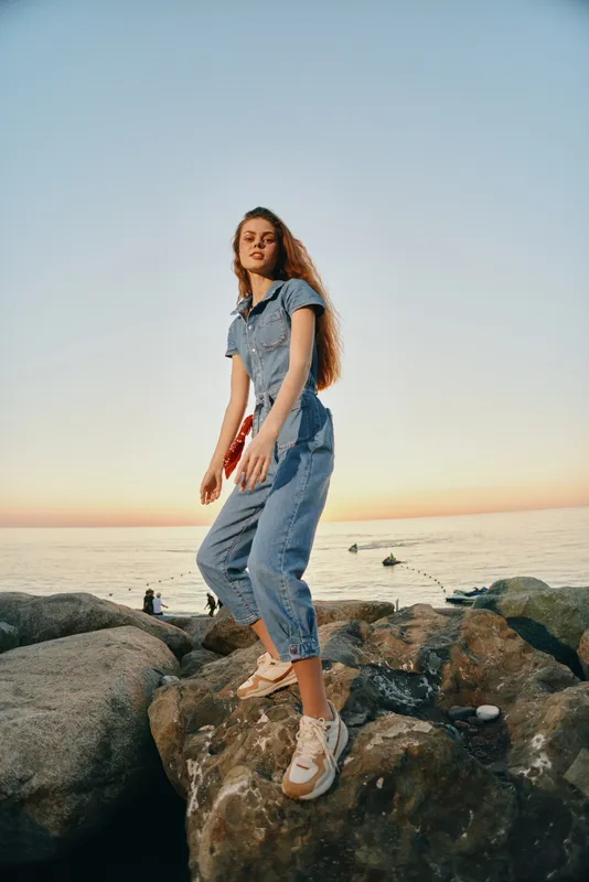 young woman standing on rocks near the sea at sunset, wearing casual denim outfit, with calm ocean and warm sky in background