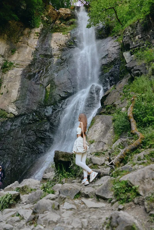 Woman hiking near a tall waterfall surrounded by rocks and lush green forest, wearing white activewear, enjoying nature outdoors on a sunny day.