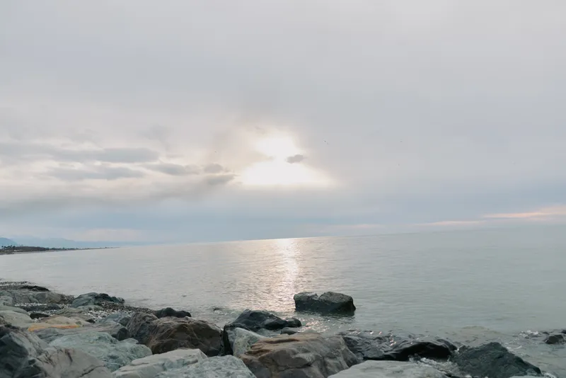 Sunset over calm sea with rocky shore in foreground under cloudy sky. Peaceful coastal landscape with soft natural light reflecting on water surface.