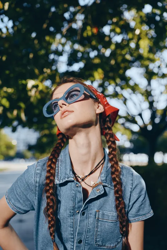 Lifestyle woman in modern boho-western denim street style with braided hair, oversized sunglasses, and a red bandana under natural sunlight, film color effect.